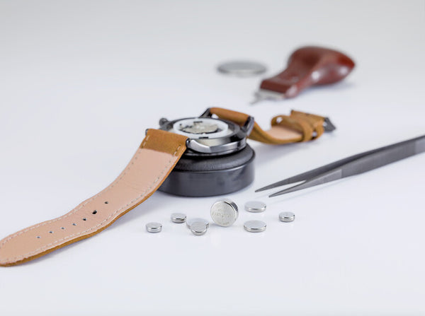 Watch case open beside a strap, tweezers and small batteries